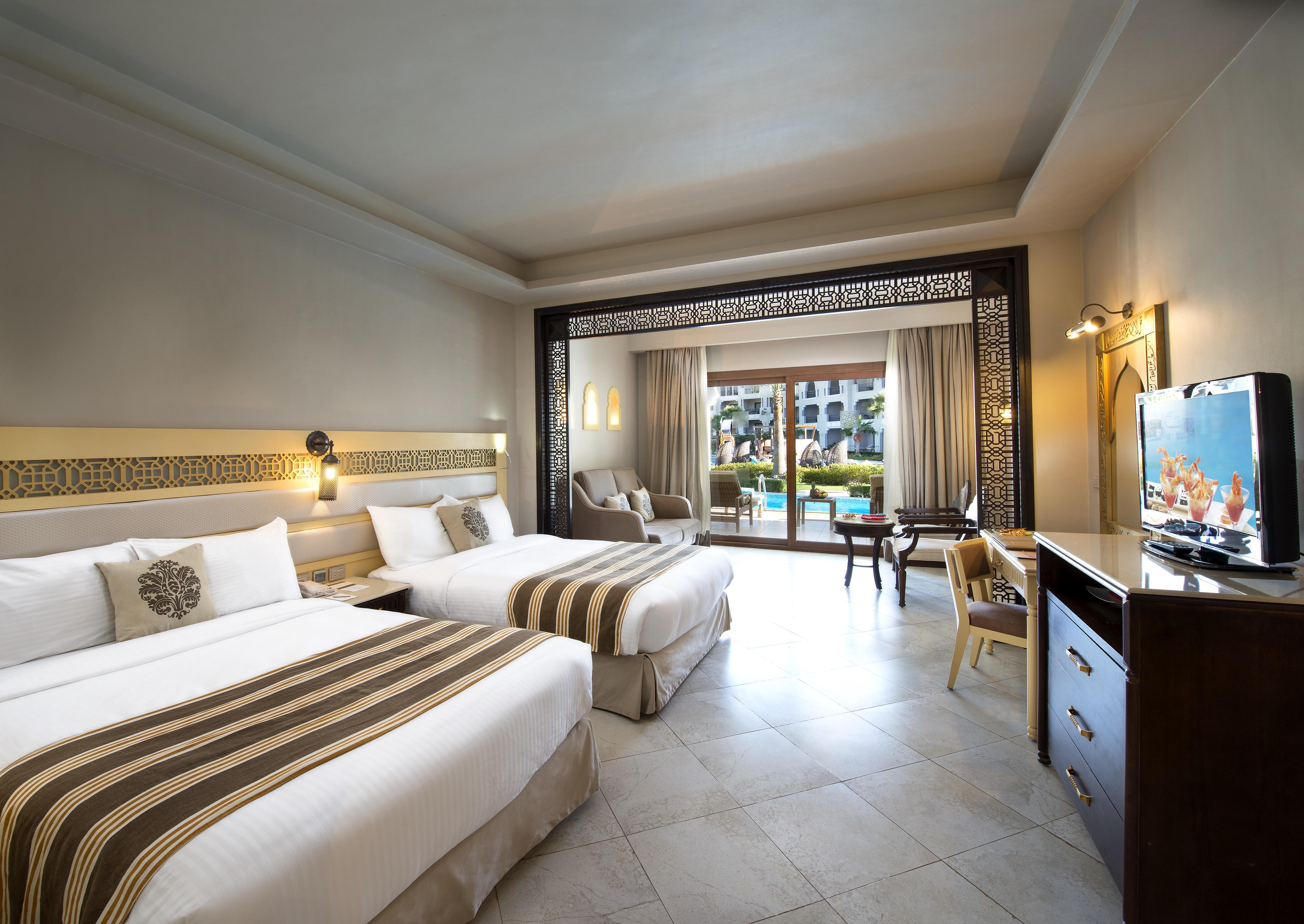 Sunrise Arabian Beach Resort Sharm el-Sheikh Exterior photo The photo shows a well-decorated hotel room. It features two beds with white linens and decorative brown striped accents. There is a large window with sheer curtains that provides a view of the outdoors, suggesting a pool area or gardens. The room in