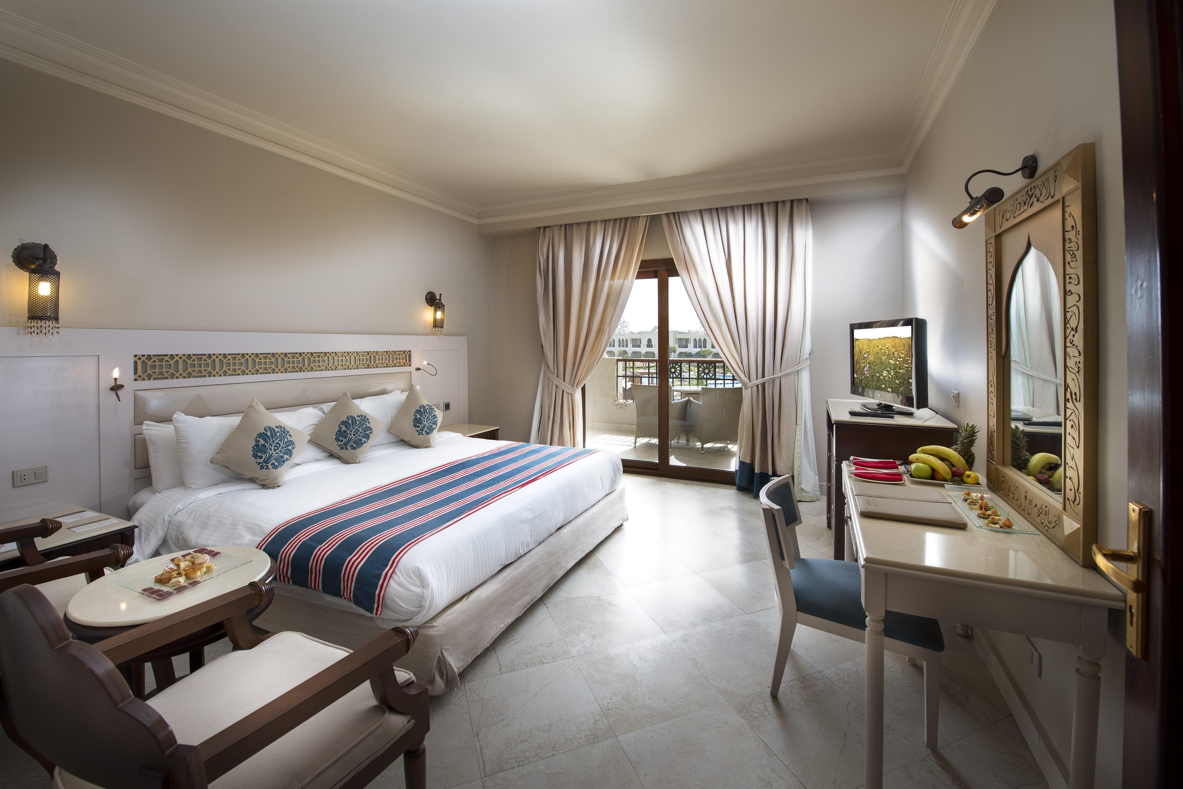 Sunrise Arabian Beach Resort Sharm el-Sheikh Exterior photo The photo depicts a hotel room that appears stylish and well-furnished. It features a large bed with decorative pillows and a colorful blanket. There is a small seating area with armchairs and a round table, as well as a work desk with a chair. A tel