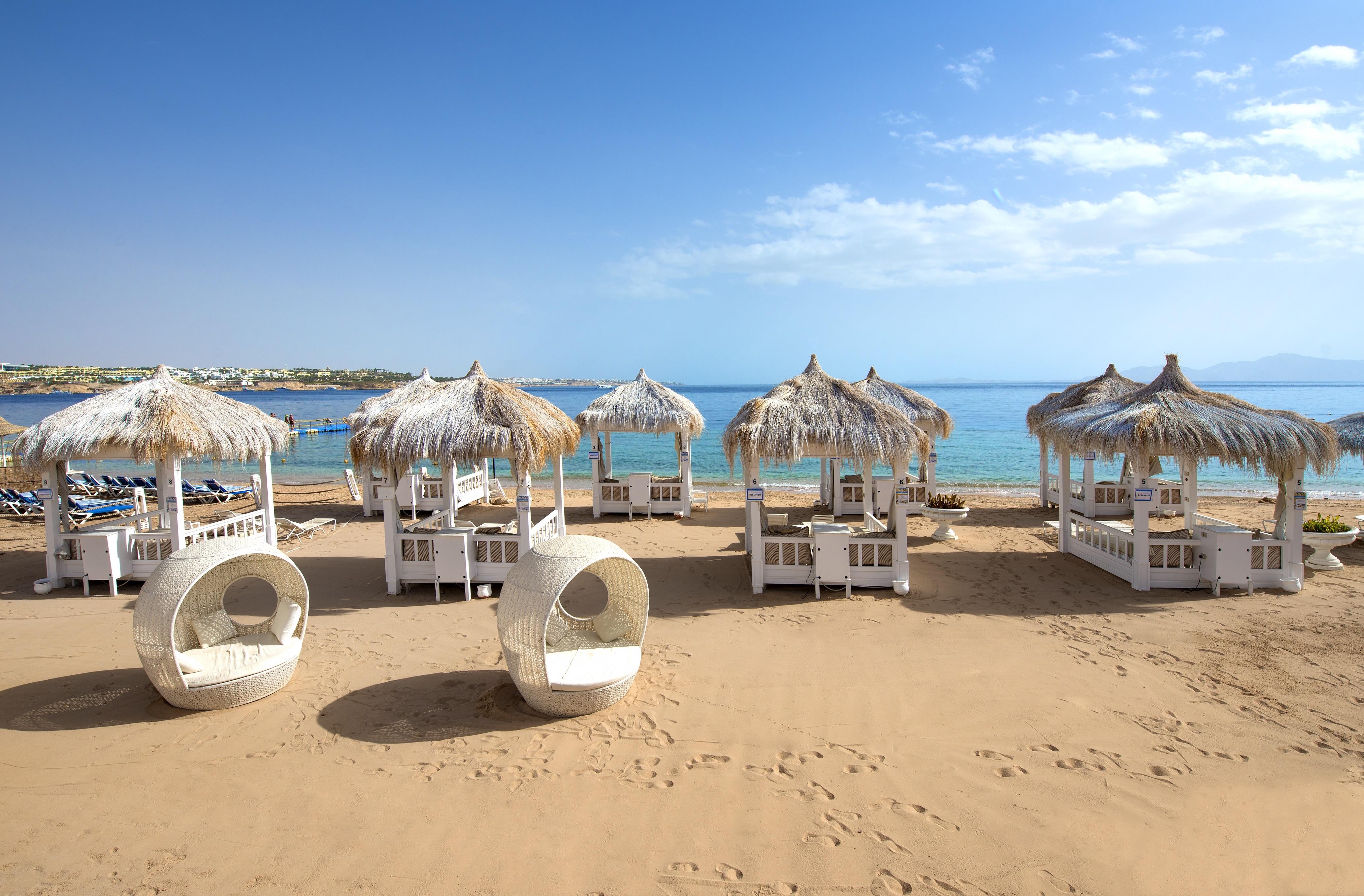 Sunrise Arabian Beach Resort Sharm el-Sheikh Exterior photo The photo shows a picturesque beach scene featuring several stylish white cabanas with thatched roofs, set on golden sand. They are arranged neatly, with some circular lounge chairs nearby. In the background, the calm blue sea stretches out under a c