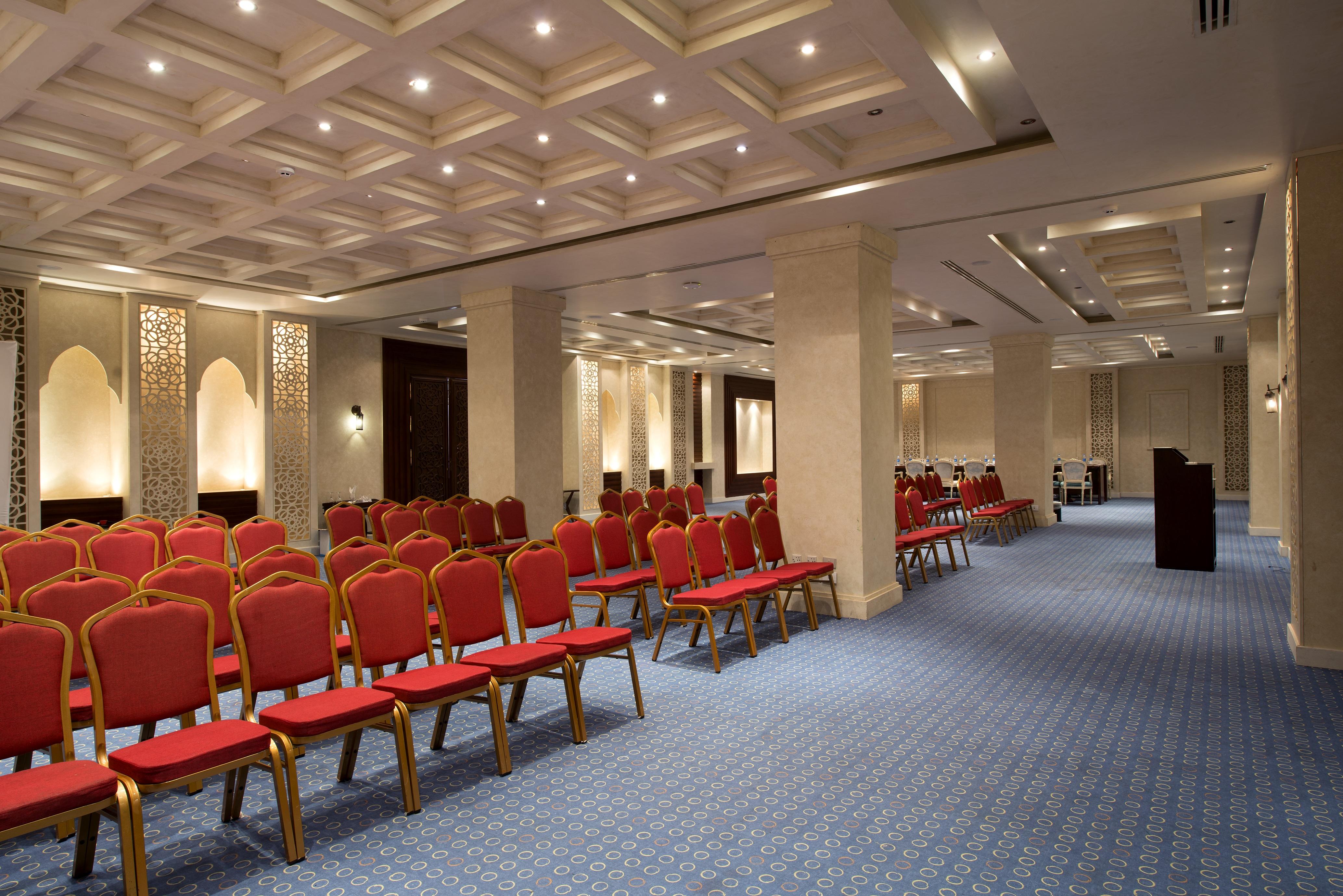 Sunrise Arabian Beach Resort Sharm el-Sheikh Exterior photo The image shows a spacious indoor venue designed for meetings or events. It features rows of red chairs arranged neatly on a blue carpeted floor. The walls are adorned with decorative panels, and the ceiling has a modern geometric design with recesse
