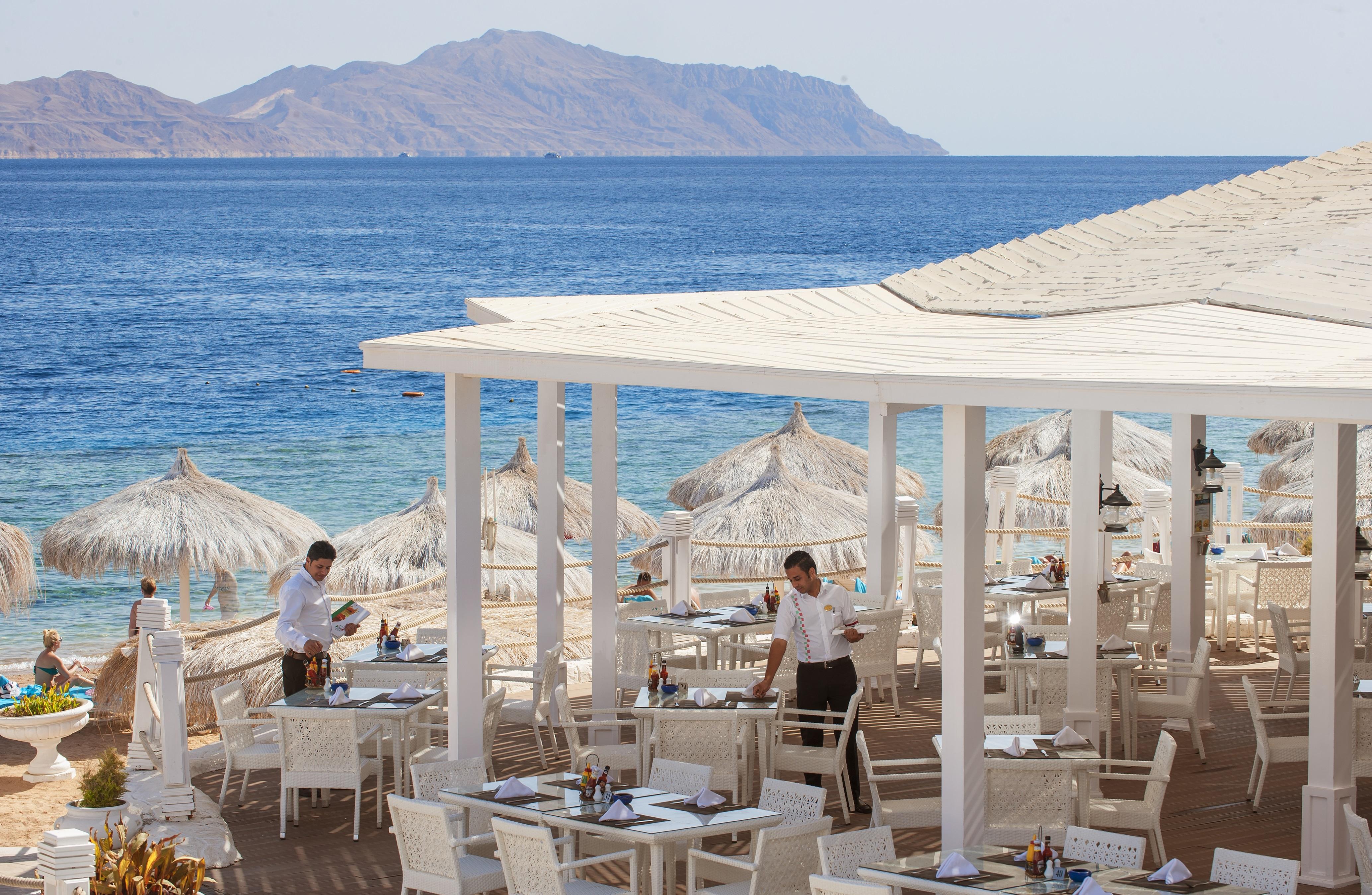 Sunrise Arabian Beach Resort Sharm el-Sheikh Exterior photo The photo depicts a seaside restaurant with a beautiful view of the ocean and distant mountains. The outdoor dining area features white tables and chairs arranged under straw umbrellas for shade. Two waitstaff members are visible, attending to the ta