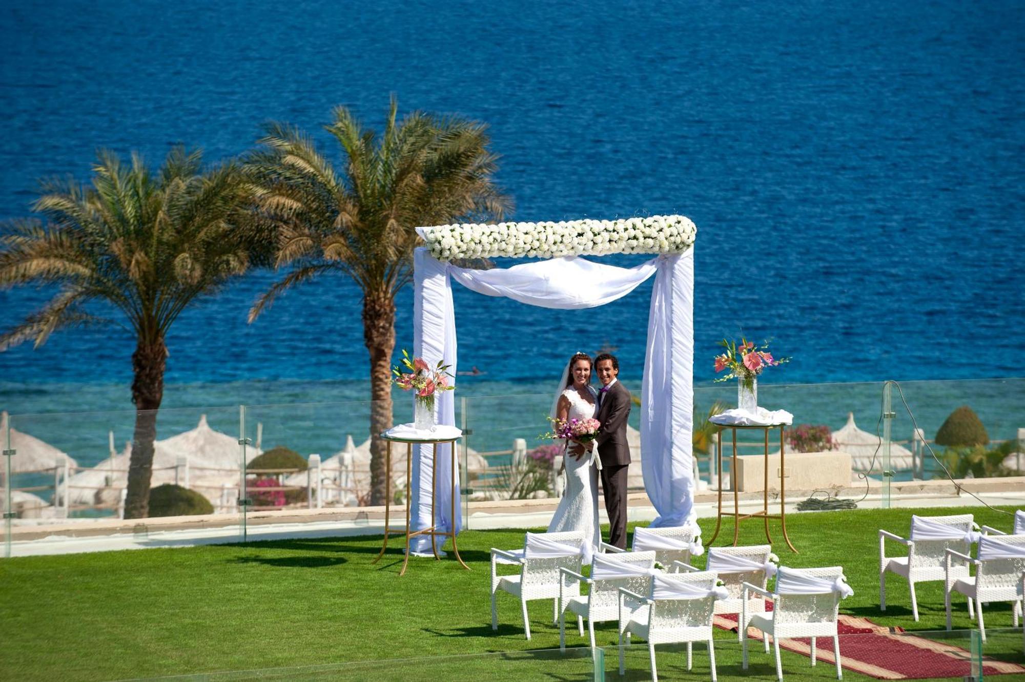 Sunrise Arabian Beach Resort Sharm el-Sheikh Exterior photo The photo depicts a wedding scene set against a beautiful seaside backdrop. In the foreground, a couple is standing underneath a decorative wedding arch adorned with white fabric and flowers. The groom is in a dark suit, while the bride is dressed in