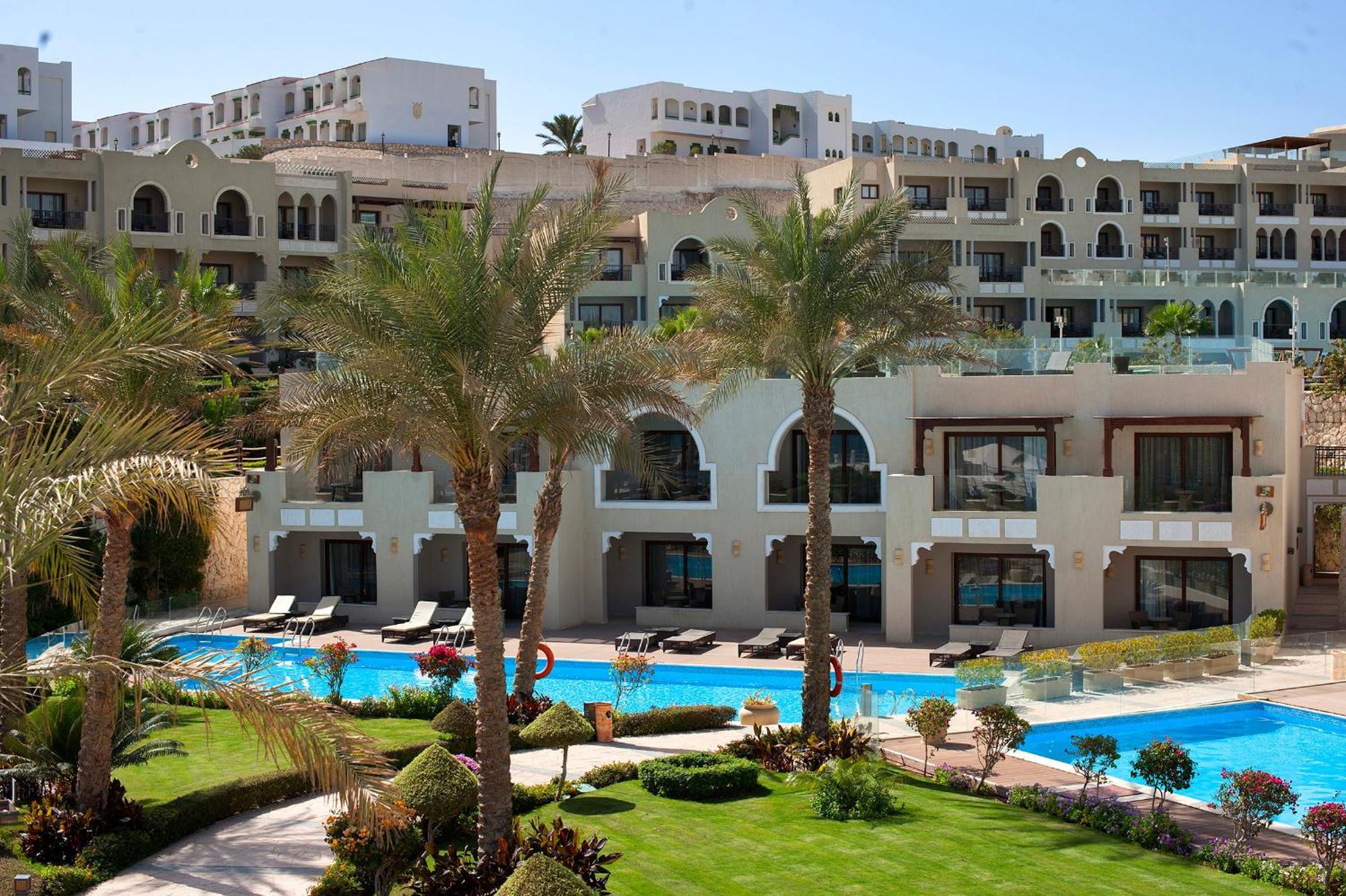 Sunrise Arabian Beach Resort Sharm el-Sheikh Exterior photo The photo depicts a luxurious resort or hotel complex. In the foreground, there is a landscaped area with manicured lawns and palm trees. A swimming pool is visible, reflecting the clear blue sky. The background features several multi-story buildings