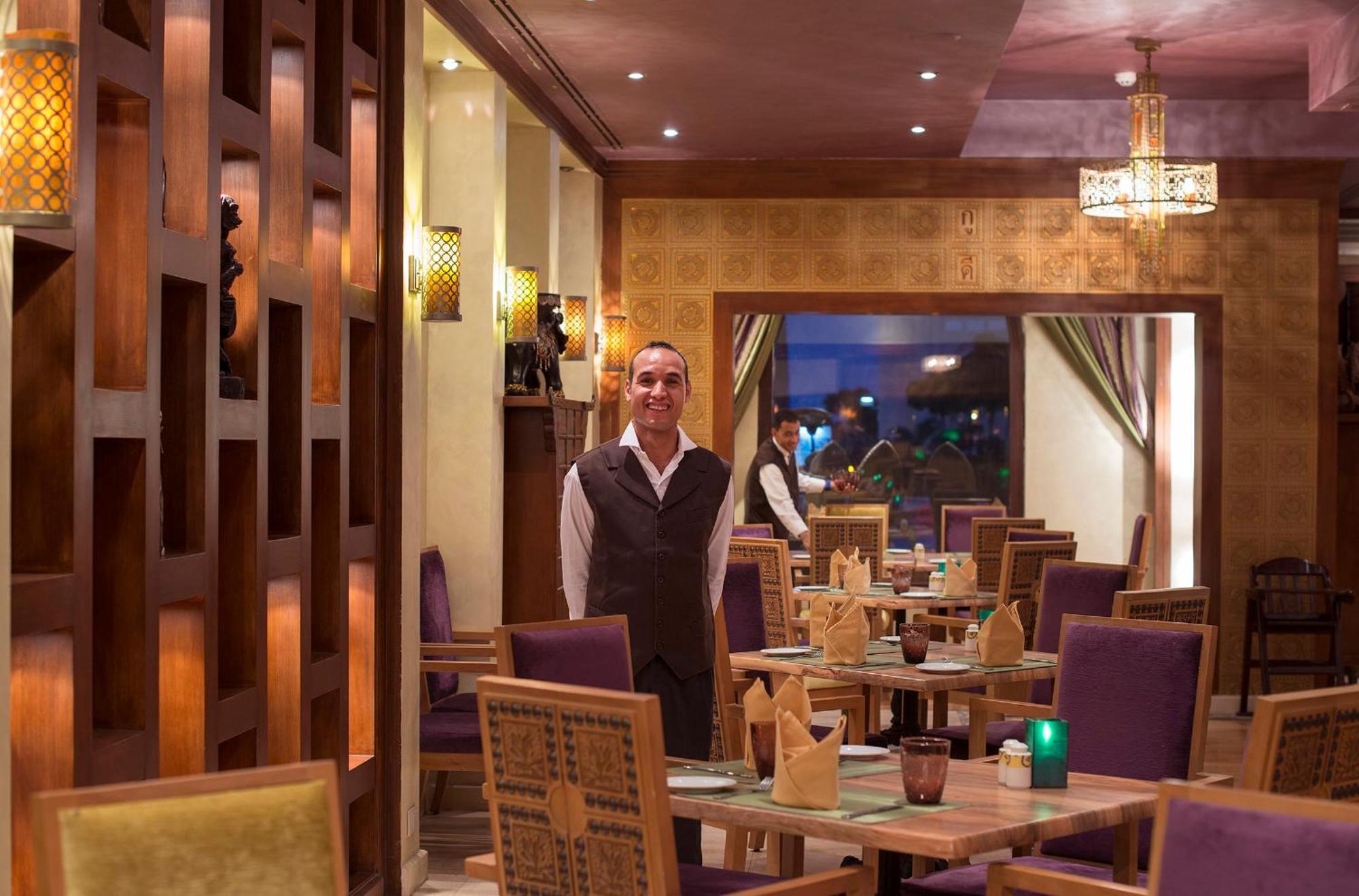 Sunrise Arabian Beach Resort Sharm el-Sheikh Exterior photo The photo depicts the interior of a restaurant featuring a staff member standing in the foreground. The restaurant has a warm and inviting ambiance, characterized by wooden accents, stylish lighting, and neatly arranged tables. The seating includes p